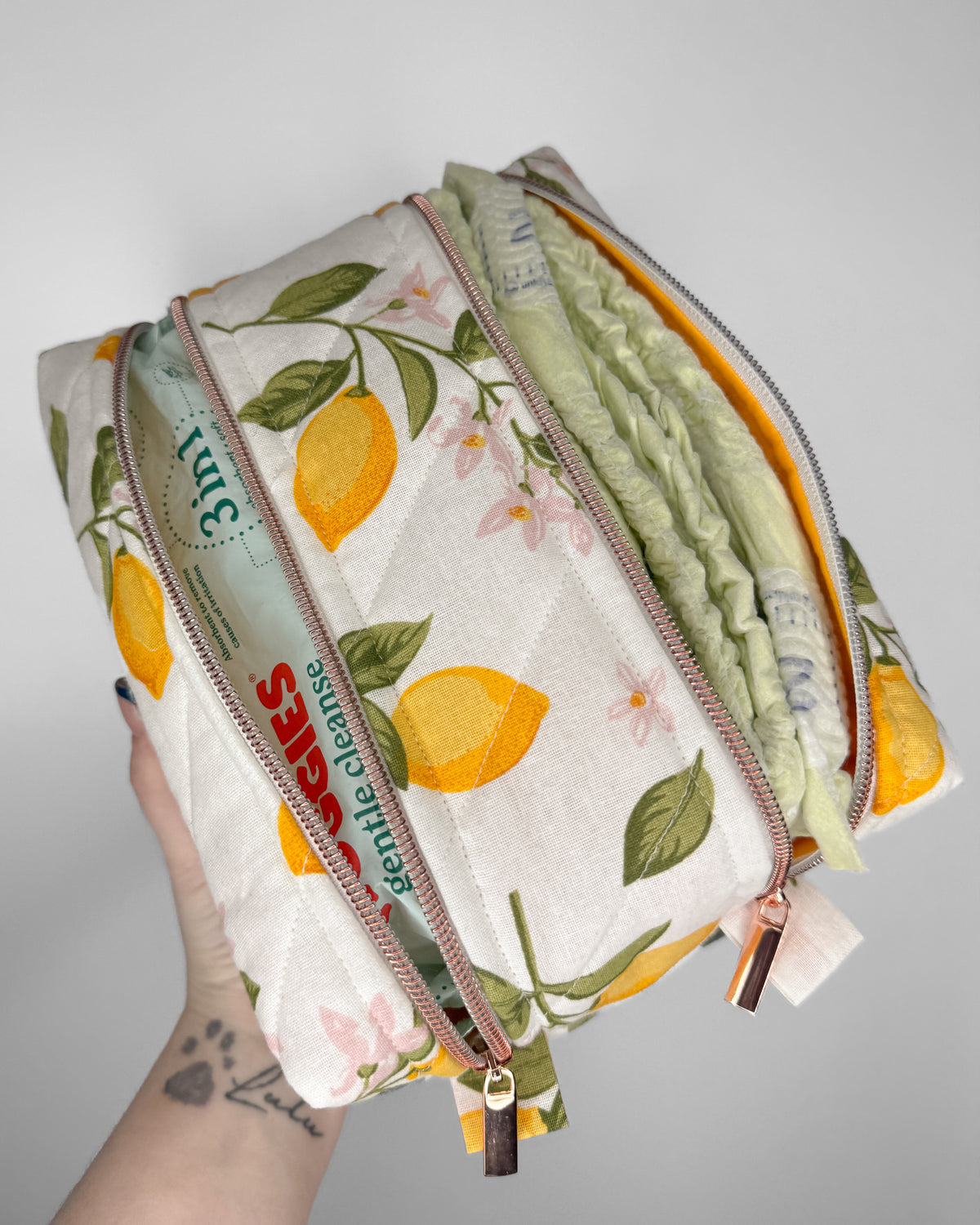 A hand holds a quilted zippered pouch with a lemon and floral print, showing a soft green reusable diaper inside. The pouch has rose gold zippers and a visible '3 in 1' label.