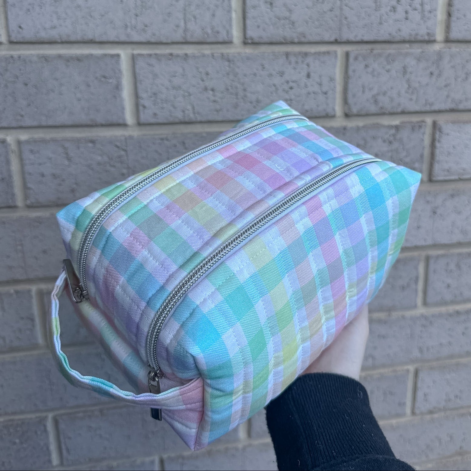 Pastel plaid bag held by a person against a brick wall.