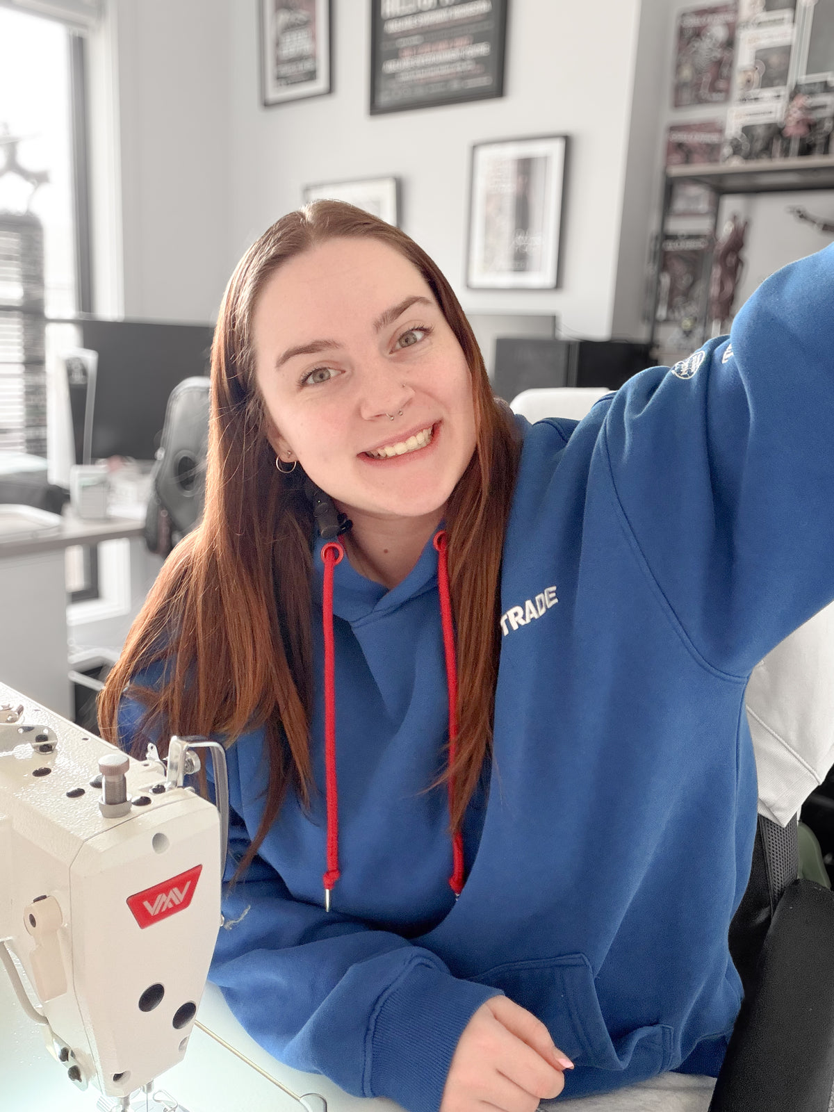 A smiling woman with long brown hair wearing a blue hoodie with red drawstrings and a white 'TRADE' logo on the chest, sitting at a sewing machine in an office setting.