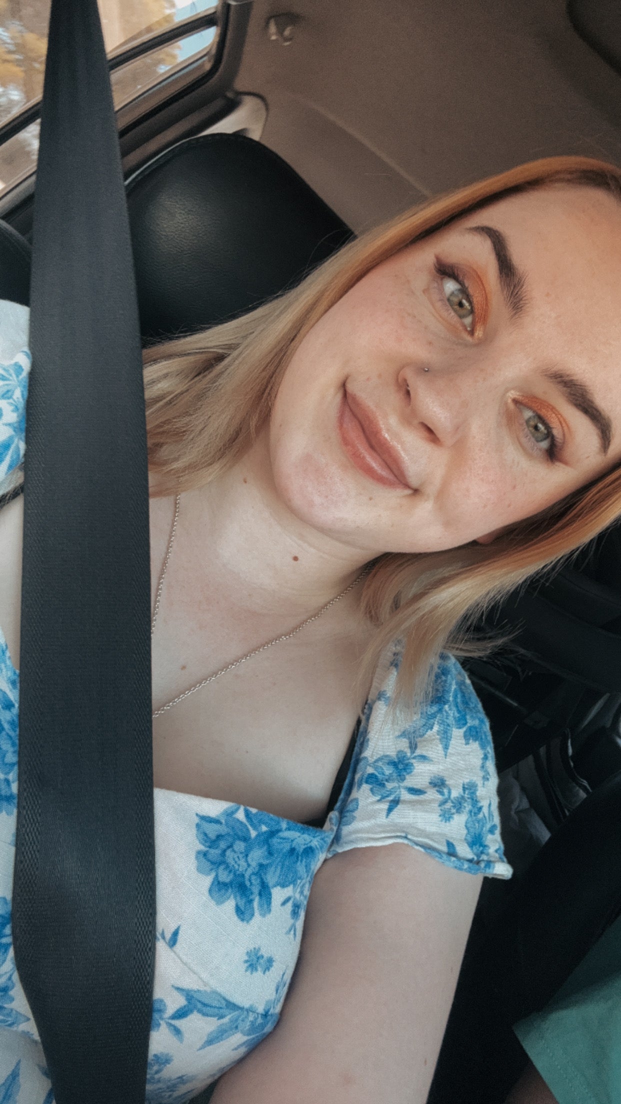 A close-up selfie of a woman in a car wearing a blue and white floral print top, with a black seatbelt across her chest.